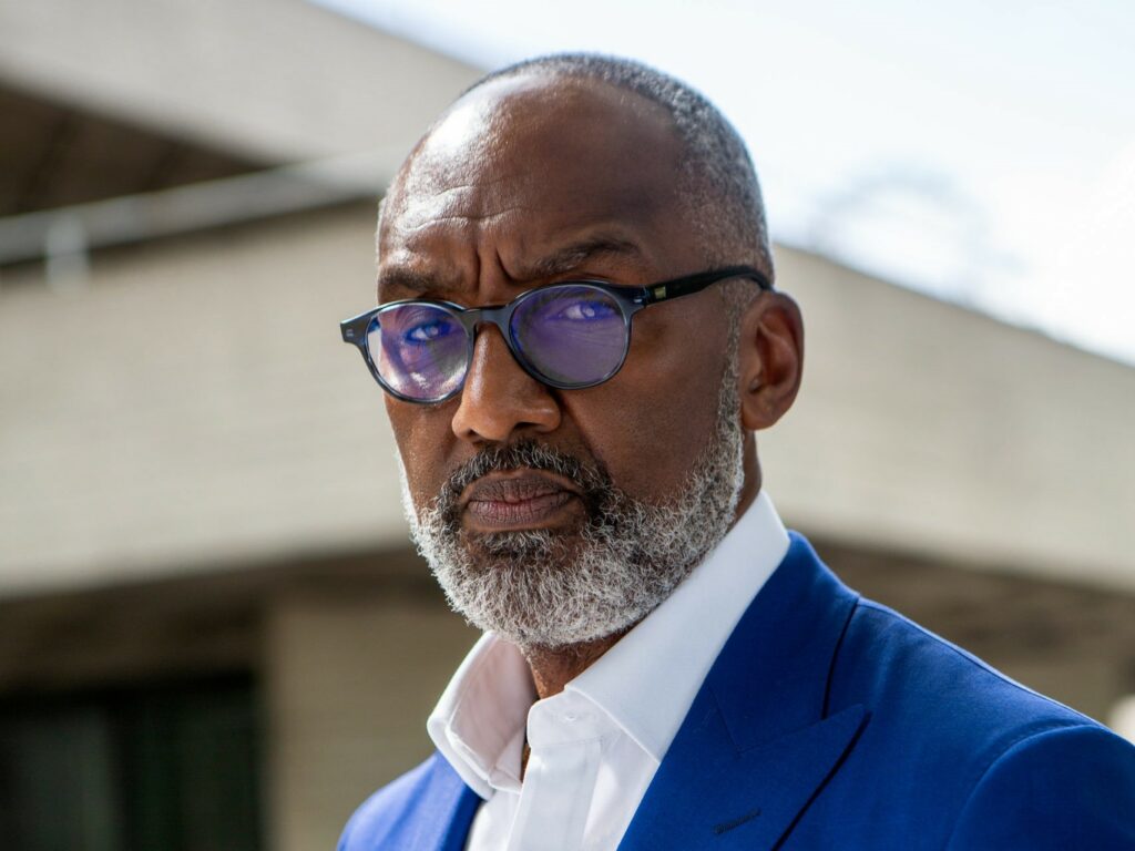 A Black person with a grey beard, blue suit, and glasses