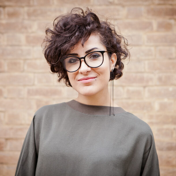 A person with a light skin tone and short curly dark brown hair, wearing glasses and a grey top